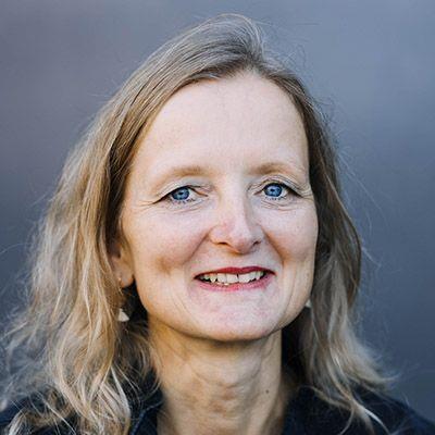 Porträt von Franziska von Oppen: eine lächelnde Frau mittleren Alters mit hellblauen Augen und schulterlangen, leicht gewellten blonden Haaren vor einem neutralen grauen Hintergrund